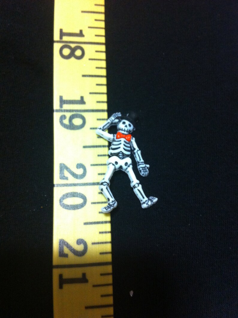May include: A white skeleton figurine wearing a black top hat and a red bow tie. The figurine is about 1 inch tall and is standing on a black surface.