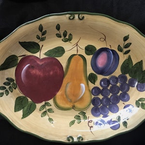 Puede incluir: Un plato de servir ovalado de cerámica con un diseño de fruta pintado a mano. El plato presenta una manzana roja, una pera amarilla, una ciruela morada y un racimo de uvas moradas. El fondo es beige claro con hojas verdes.