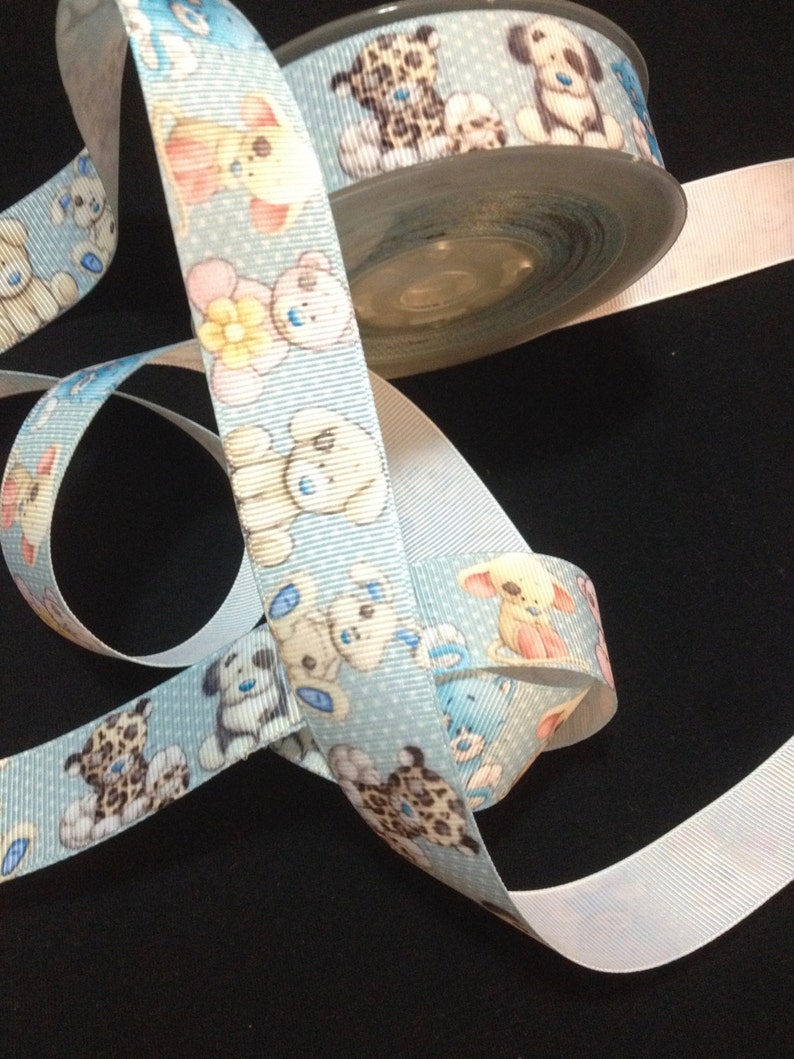 May include: A blue ribbon with a white polka dot pattern and a repeating design of teddy bears and a leopard print animal. The ribbon is rolled up on a spool.