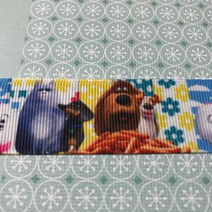 May include: A colorful ribbon with a cartoon image of a group of dogs and cats. The ribbon features a brown dog, a white dog, a gray cat, and a dachshund. The ribbon has a yellow background with blue and green flowers.