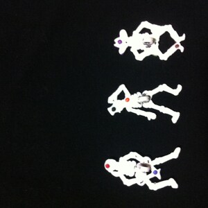 May include: Three white plastic skeleton figurines with colorful accents. The skeletons are posed in different positions, with one sitting, one standing, and one lying down.