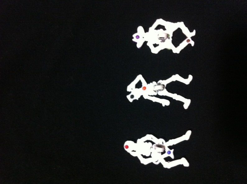 May include: Three white plastic skeleton figurines with colorful accents. The skeletons are posed in different positions, with one sitting, one standing, and one lying down.