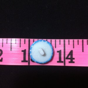 May include: A white button with a blue and white polka dot trim, measuring 1 inch in diameter. The button is on a pink measuring tape.