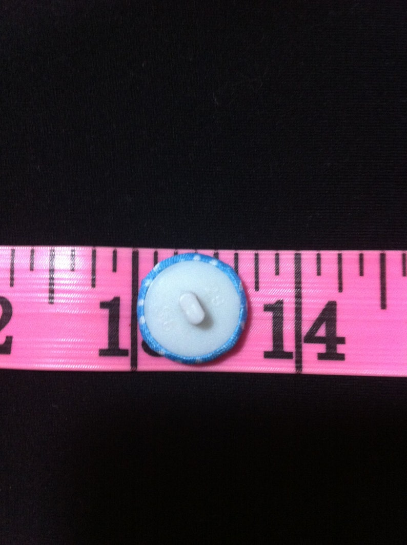 May include: A white button with a blue and white polka dot trim, measuring 1 inch in diameter. The button is on a pink measuring tape.