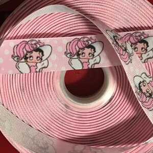 以下が含まれることがあります： 白い背景にピンクのリボンが巻かれており、帽子をかぶった漫画の女の子が繰り返し描かれています。リボンは巻かれており、そのパターンはロールの外側に表示されています。