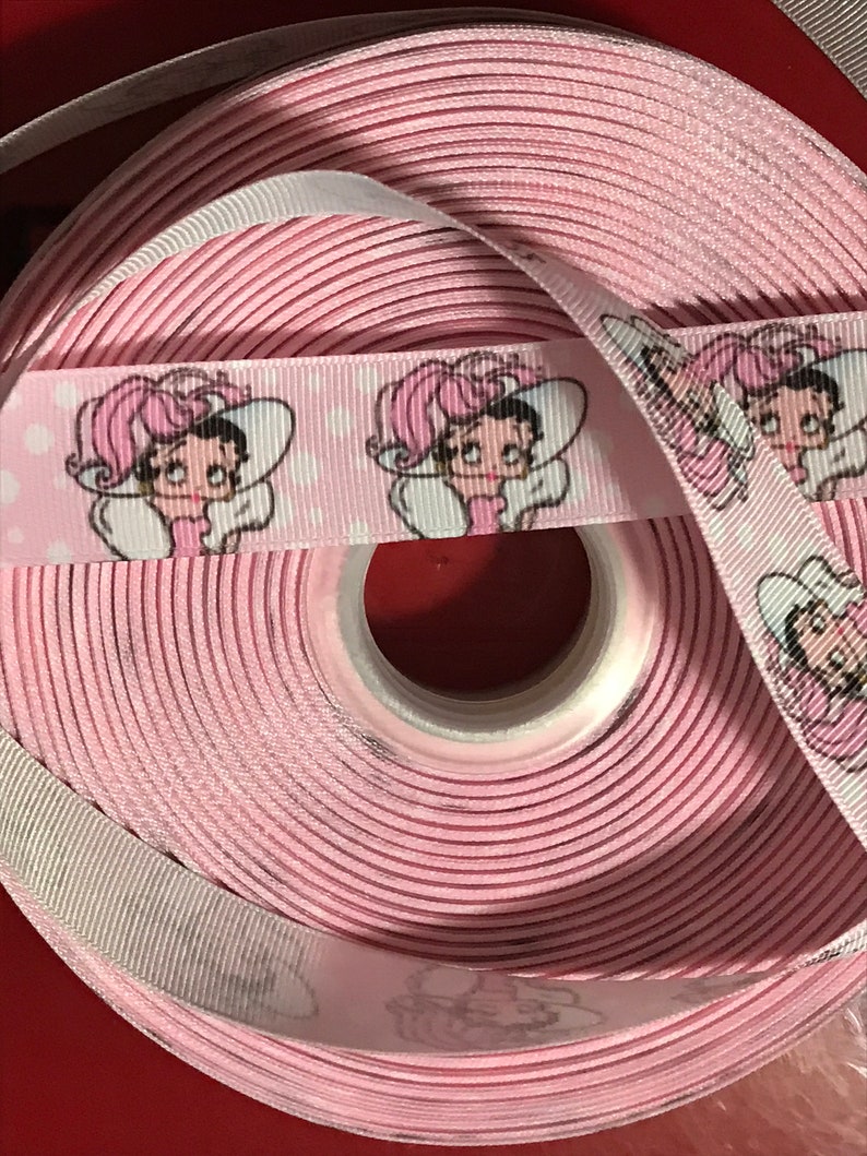 以下が含まれることがあります： 白い背景にピンクのリボンが巻かれており、帽子をかぶった漫画の女の子が繰り返し描かれています。リボンは巻かれており、そのパターンはロールの外側に表示されています。