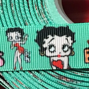 May include: A roll of turquoise ribbon with a white background and a repeated pattern of a cartoon character with black hair and red lips. The character is wearing a red dress and holding a heart.