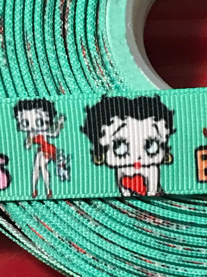 May include: A roll of turquoise ribbon with a white background and a repeated pattern of a cartoon character with black hair and red lips. The character is wearing a red dress and holding a heart.