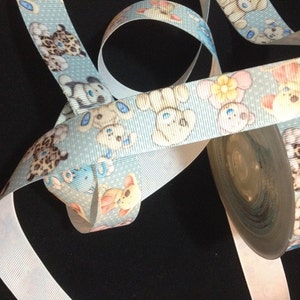 May include: A roll of blue ribbon with a white polka dot pattern and a repeating design of cartoon teddy bears. The ribbon is on a white spool.
