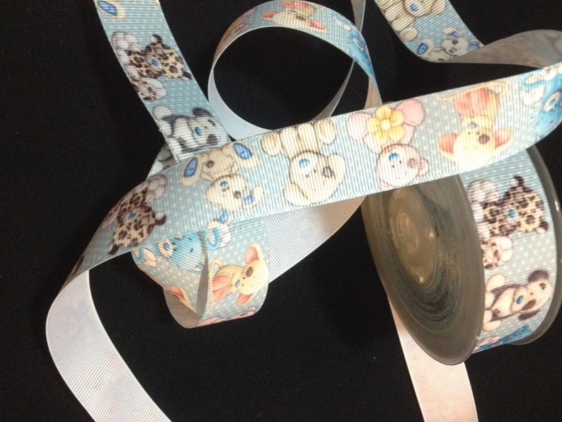 May include: A roll of blue ribbon with a white polka dot pattern and a repeating design of cartoon teddy bears. The ribbon is on a white spool.
