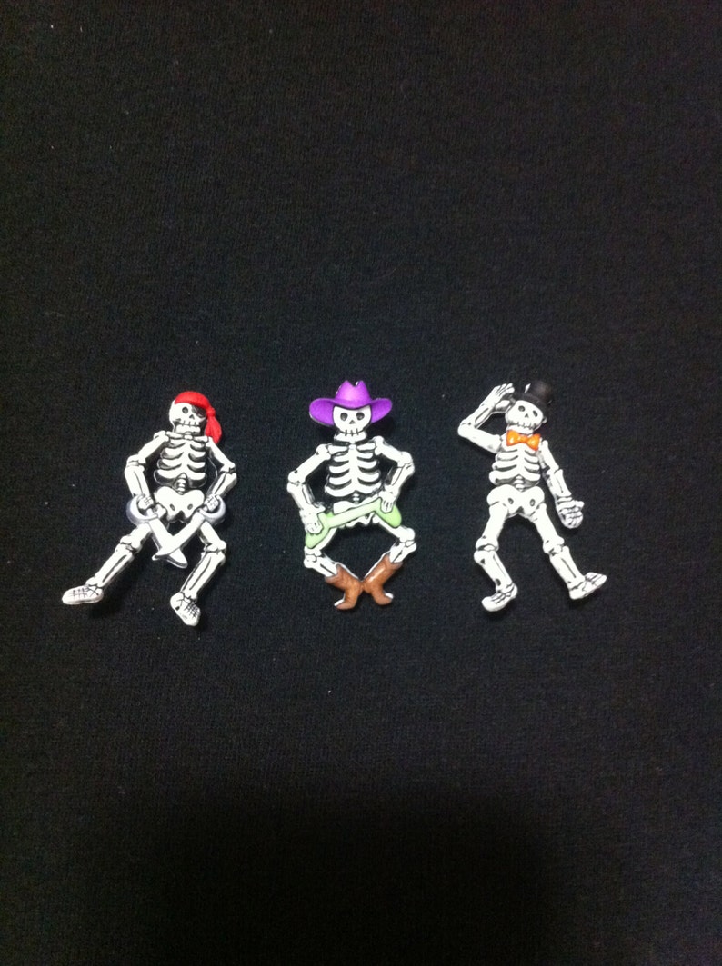 May include: Three skeleton figurines with different accessories. The skeleton on the left is wearing a red bandana and holding a sword. The skeleton in the middle is wearing a purple cowboy hat and green pants. The skeleton on the right is wearing a black top hat and a bow tie.