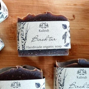 Può includere: Barrette di sapone biologico fatte a mano di colore marrone scuro con una superficie strutturata. Ogni saponetta è avvolta con un'etichetta di carta con la scritta "Birchtar" e un'illustrazione di betulla. I saponi sono disposti su una superficie di legno.