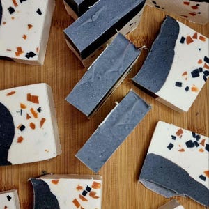 Puede incluir: Barras de jabón artesanal con un diseño moderno. Los jabones presentan una combinación de colores blanco, gris, negro y naranja. Algunas barras tienen un patrón tipo terrazo, mientras que otras tienen una capa superior gris sólida. Los jabones están sobre una superficie de madera.