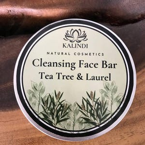 May include: A round, silver-rimmed tin of Kalindi Natural Cosmetics Cleansing Face Bar. The label features a lotus flower logo, the words "Cleansing Face Bar Tea Tree & Laurel," and botanical illustrations in green and white.