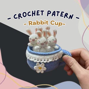 Op de afbeelding: Een gehaakte blauwe theekop met witte details, met meerdere witte gehaakte konijnen met roze accenten. De theekop heeft een bloem en de tekst "CROCHET PATTERN - Rabbit Cup-".