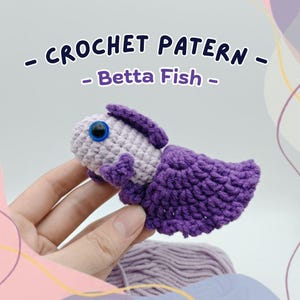 Può includere: Un pesce Betta all'uncinetto nei toni del viola e del grigio chiaro. Il pesce ha una grande coda strutturata e una piccola pinna sulla parte superiore. Il testo "- CROCHET PATTERN - Betta Fish -" è visualizzato sopra il pesce.
