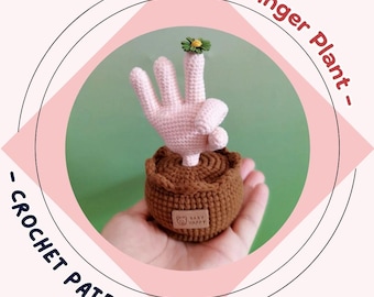 May include: A crocheted finger plant in a brown pot. The plant is a light pink hand with a green and yellow flower on the index finger. The pot has a small tag that says "Baby Happy". The background is green.