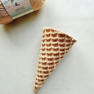 Może przedstawiać: Szydełkowy rożek do lod&oacute;w z wzorem wafelka, w odcieniach beżu i jasnego brązu. W tle znajduje się motek beżowej przędzy. Stożek jest ręcznie robiony i wydaje się być częścią projektu rzemieślniczego.