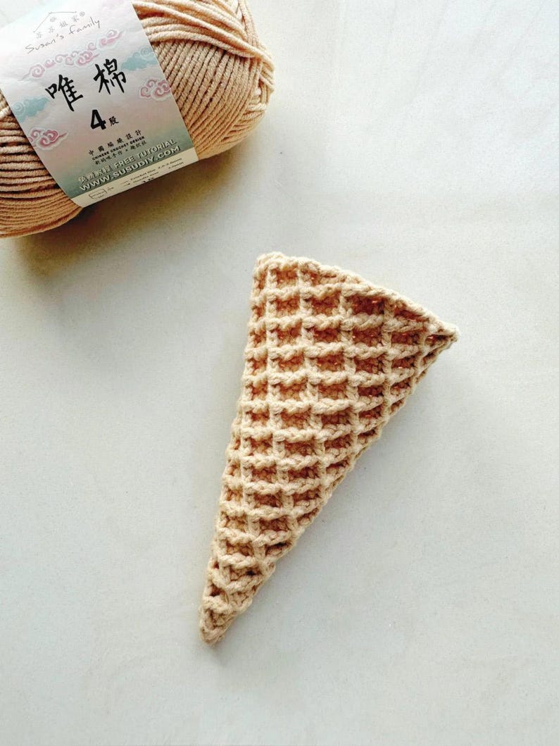 Może przedstawiać: Szydełkowy rożek do lod&oacute;w z wzorem wafelka, w odcieniach beżu i jasnego brązu. W tle znajduje się motek beżowej przędzy. Stożek jest ręcznie robiony i wydaje się być częścią projektu rzemieślniczego.