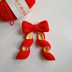 Puede incluir: Un lazo rojo hecho a mano con ganchillo, con detalles amarillos y espirales. Junto al lazo hay un ovillo de lana roja con texto chino en la etiqueta. La lana es de color rojo brillante, ideal para proyectos de manualidades.