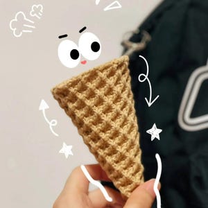 Może przedstawiać: Szydełkowy rożek do lod&oacute;w w jasnobrązowym kolorze. Stożek ma wz&oacute;r wafelka i jest trzymany przez dłoń. Fantazyjne bazgroły oczu, pary, strzałek i gwiazd otaczają rożek.