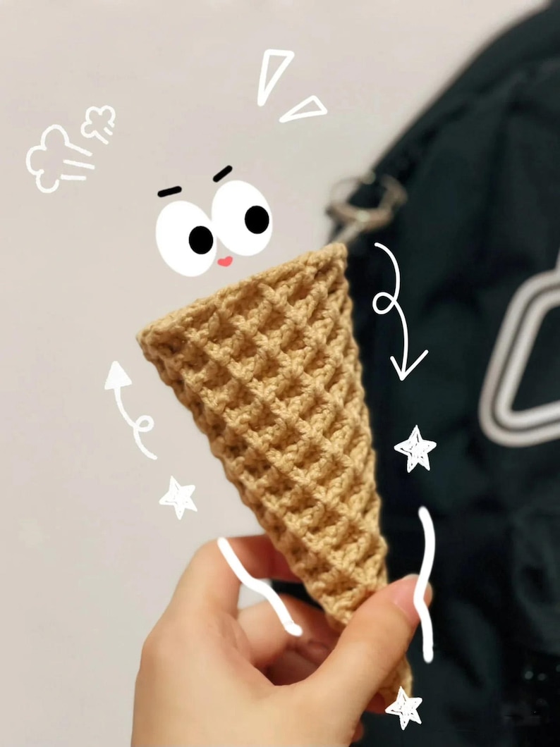 Może przedstawiać: Szydełkowy rożek do lod&oacute;w w jasnobrązowym kolorze. Stożek ma wz&oacute;r wafelka i jest trzymany przez dłoń. Fantazyjne bazgroły oczu, pary, strzałek i gwiazd otaczają rożek.