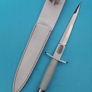 May include: A silver dagger with a white ribbed handle and a matching white leather sheath. The dagger has a pointed blade and a crossguard. The word "CASE" is printed on the blade. The sheath has a strap with a snap closure.