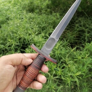 Puede incluir: Una daga con una hoja plateada de doble filo y un mango envuelto en cuero marrón. La guarda es roja y la palabra "CASE" está grabada en la hoja. La daga se sostiene sobre un fondo de follaje verde.