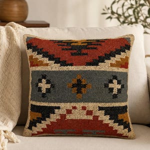 May include: A square throw pillow featuring a woven texture with a geometric pattern in shades of rust red, charcoal black, mustard yellow, and cream. The design includes horizontal bands with stepped motifs and central cross-shaped accents.