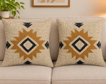 Puede incluir: Dos cojines cuadrados con una base de tela beige texturizada. Cada cojín muestra un patrón geométrico central de diamante en amarillo mostaza y negro, con bordes dentados en forma de estrella que se extienden horizontal y verticalmente.