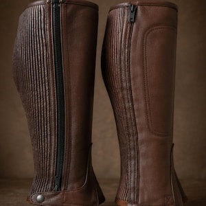 May include: A pair of brown leather equestrian half chaps featuring a vertical ribbed texture on the outer side, a full-length black zipper closure, and a reinforced panel on the inner calf. Each chap includes a snap button at the bottom hem.