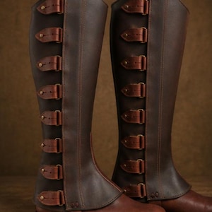 May include: A pair of knee-high dark brown leather boots featuring a vertical panel on the outer side secured with seven individual buckled straps. The boots have a smooth finish, a rounded toe, and a low heel, with visible stitching along the seams.