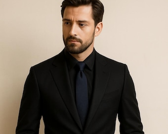 May include: A black suit with a matching black shirt and a navy blue tie. The suit has a two-button closure and a notched lapel. The person is wearing a black belt. The background is a solid, neutral color.
