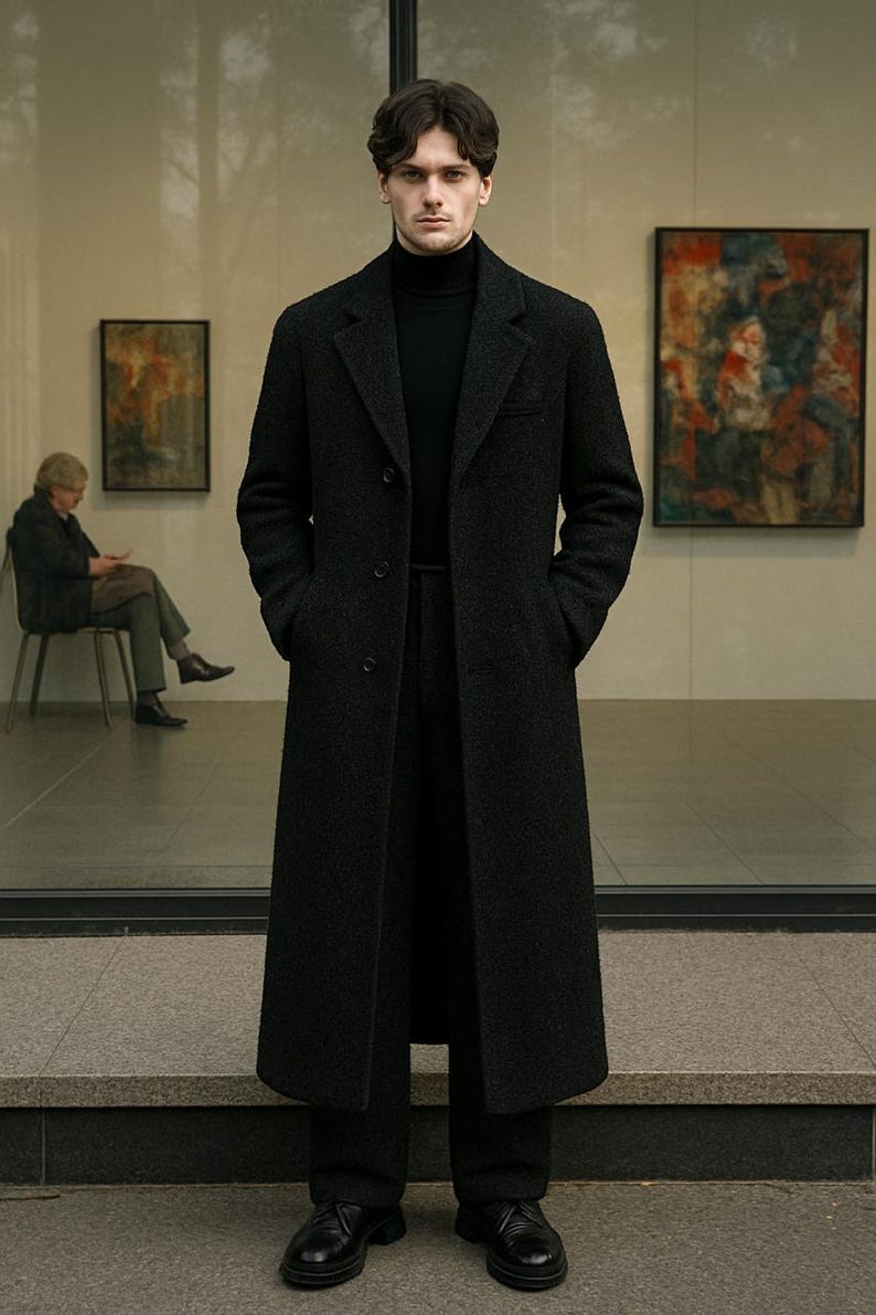 May include: A man wearing a black wool coat and matching pants. The coat is long, with a classic lapel and pockets. He is also wearing a black turtleneck. The background includes a building with large windows and artwork.