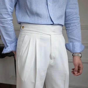 May include: White pleated trousers with a high waist and a wrap-around design. The trousers are paired with a light blue long-sleeve shirt. A gold button detail is visible on the waistband.