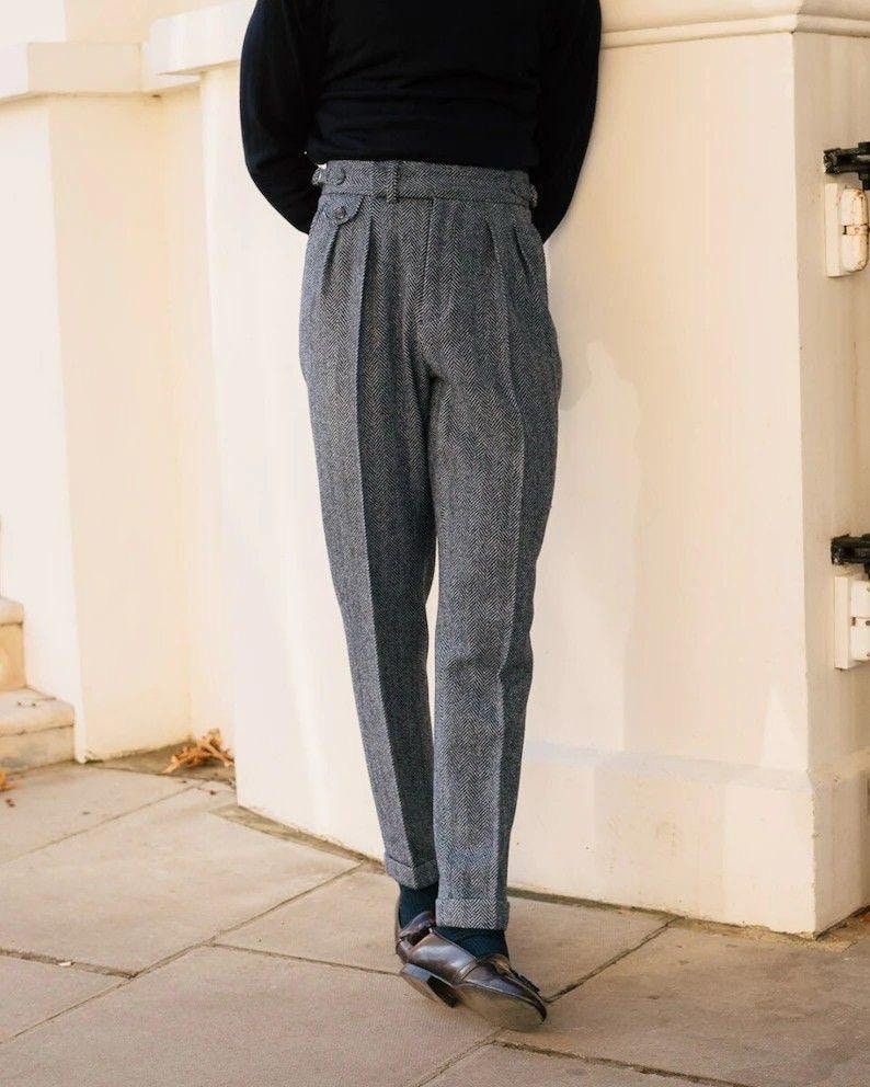 May include: Gray herringbone trousers with a high waist and pleated front. The pants have a tailored fit and are paired with dark shoes. The fabric has a subtle texture, and the overall style is classic and refined.