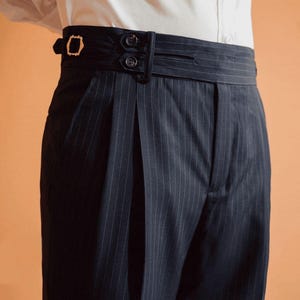 May include: Navy blue pinstripe trousers with a high-waisted design. The trousers feature a wide waistband with button and buckle details. The fabric has vertical white stripes. The trousers are part of a formal wear collection.