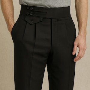 May include: Black high-waisted trousers with a wide waistband and pleats. The trousers feature a button closure and a pocket detail. The model is wearing a gray t-shirt. The trousers are a classic style.
