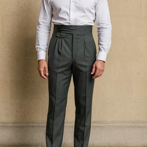 May include: A man wearing a white dress shirt and dark gray high-waisted trousers. The trousers have pleats and a buttoned closure. Brown leather dress shoes complete the outfit. The ensemble suggests formal or business attire.