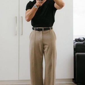 Può includere: Una persona indossa una maglietta nera, pantaloni beige e scarpe nere. La persona tiene in mano una macchina fotografica. I pantaloni hanno un design a gamba larga e sono abbinati a una cintura nera. Un orologio d'argento è visibile al polso.