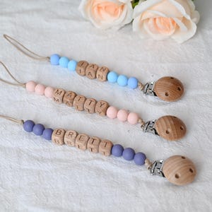 May include: Three personalized baby pacifier clips with wooden beads spelling out names. The clips feature light blue, pink, and purple beads, with wooden letters and a wooden clip. The background includes white fabric and peach-colored roses.