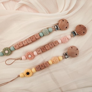 May include: Three personalized baby pacifier clips with wooden beads and floral accents. Each clip features a different name spelled out in wooden letter blocks. The clips have a variety of pastel colors, including blue, pink, and yellow.