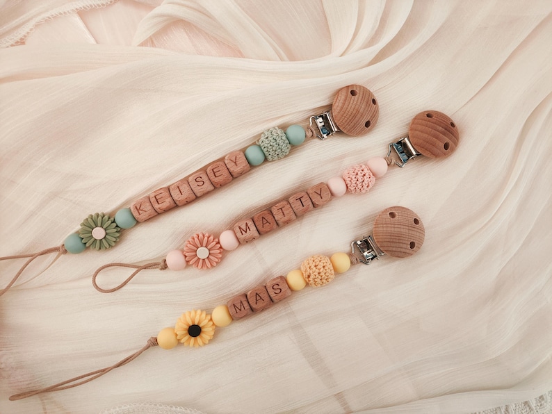 May include: Three personalized baby pacifier clips with wooden beads and floral accents. Each clip features a different name spelled out in wooden letter blocks. The clips have a variety of pastel colors, including blue, pink, and yellow.