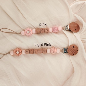 May include: Two baby pacifier clips in pink and light pink. Each clip features wooden beads spelling out a name, a flower-shaped bead, and a wooden clip. The background is a soft, light-colored fabric.