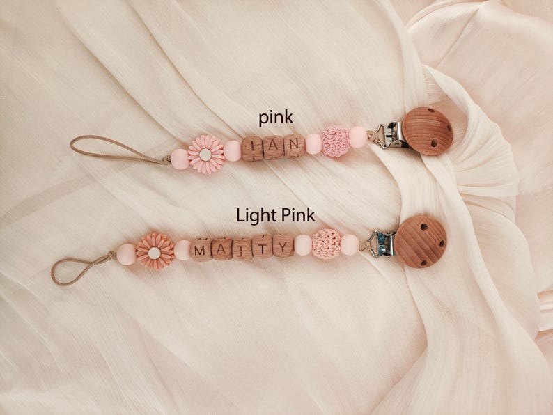 May include: Two baby pacifier clips in pink and light pink. Each clip features wooden beads spelling out a name, a flower-shaped bead, and a wooden clip. The background is a soft, light-colored fabric.