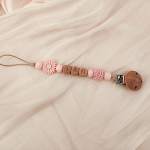 May include: A baby pacifier clip with the name "HAN" spelled out in wooden letter beads. The clip features pink beads, a pink flower, and a wooden pacifier holder. The clip is on a light pink fabric.