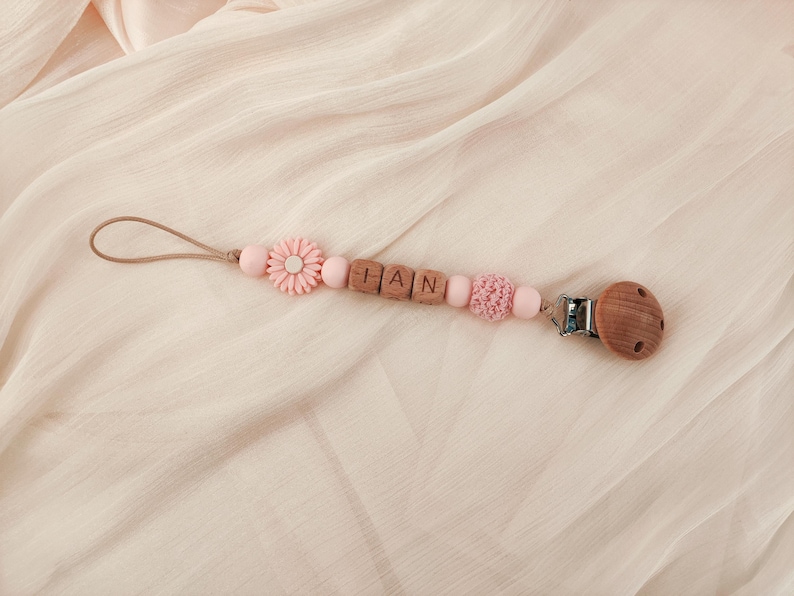 May include: A baby pacifier clip with the name "HAN" spelled out in wooden letter beads. The clip features pink beads, a pink flower, and a wooden pacifier holder. The clip is on a light pink fabric.