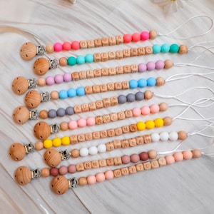 May include: A collection of personalized baby pacifier clips with wooden beads spelling out names. Each clip features a wooden clasp and a string for attachment. The beads are in various colors, including pink, blue, and yellow.