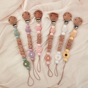 May include: Six personalized baby pacifier clips with wooden beads and names spelled out in wooden letter blocks. Each clip features a different color scheme, including green, pink, purple, white, and yellow, with decorative flower accents and wooden clips.