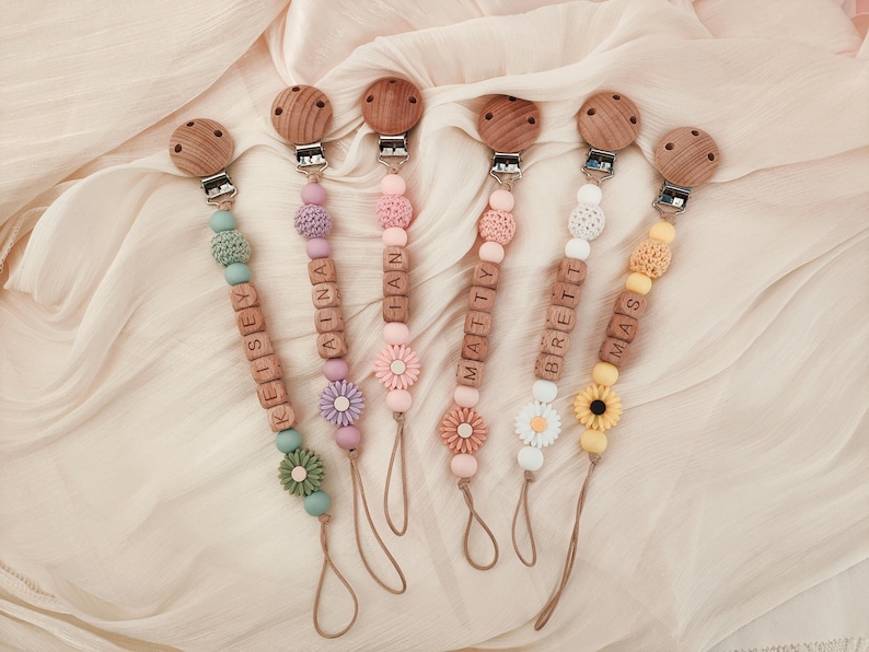 May include: Six personalized baby pacifier clips with wooden beads and names spelled out in wooden letter blocks. Each clip features a different color scheme, including green, pink, purple, white, and yellow, with decorative flower accents and wooden clips.